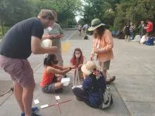 Group of six people engaged in teacher workshop activity on a paved pathway surrounded by trees; some are seated on the ground while others are standing.